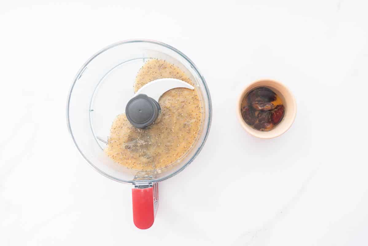 A food processor bowl with apple sauce and chia seeds on a bench next to a small dish containing soaking dates.