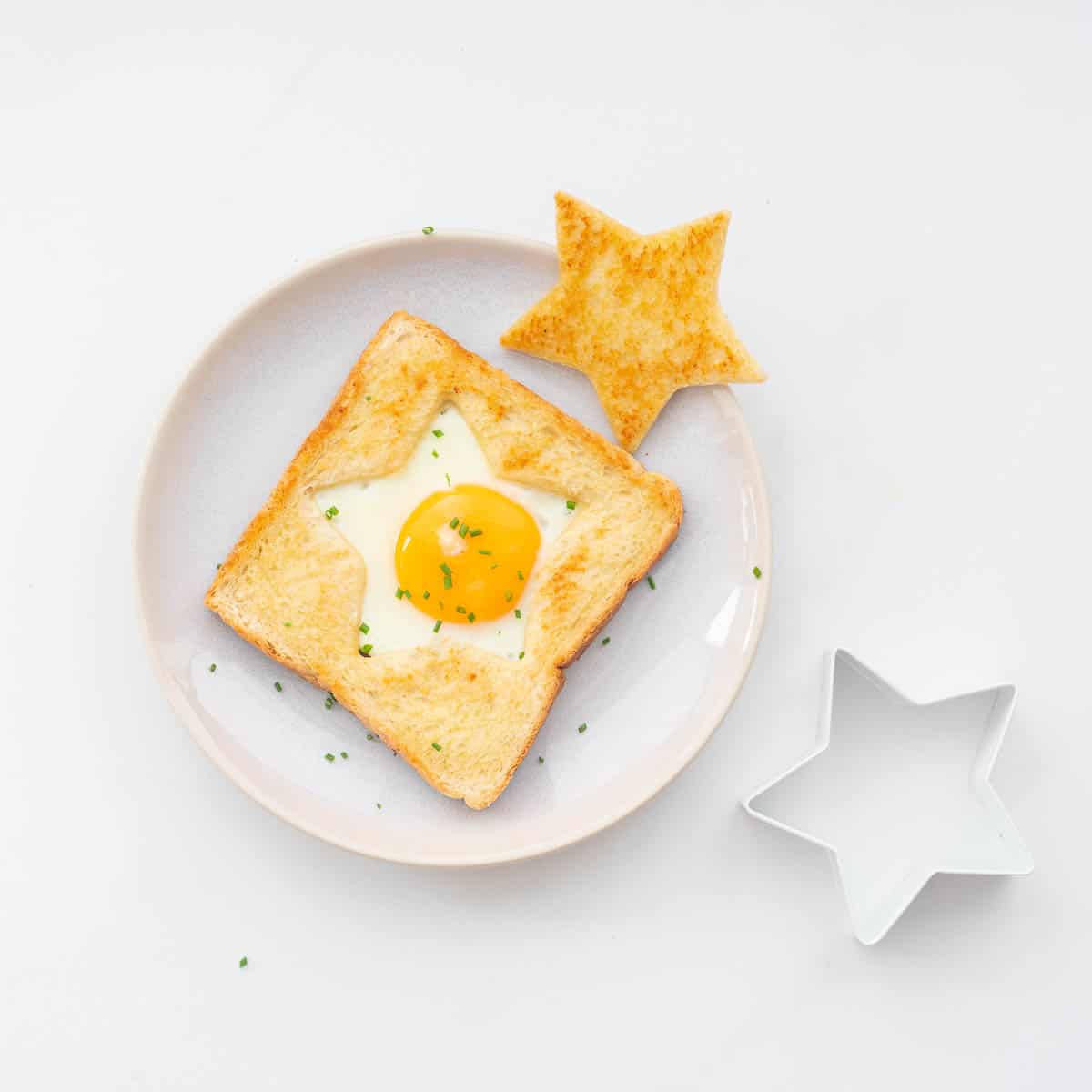 An egg cooked into the star shaped hole of a piece of toast on a plate with a golden star piece of toast to the side.