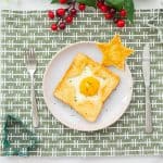 An egg cooked into the star shaped hole of a piece of toast on a plate with a golden star piece of toast to the side, sitting on.a green patterned placemat with a white green and red christmas decoration centrepiece above the placemat.