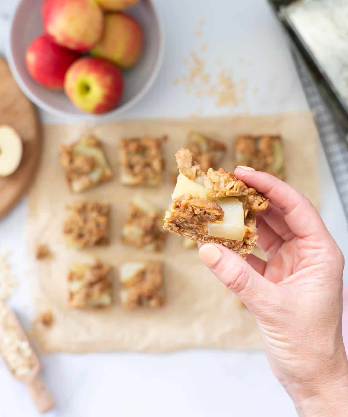 A piece of a apple crumble slice being held above a tray of apple crumble bars.