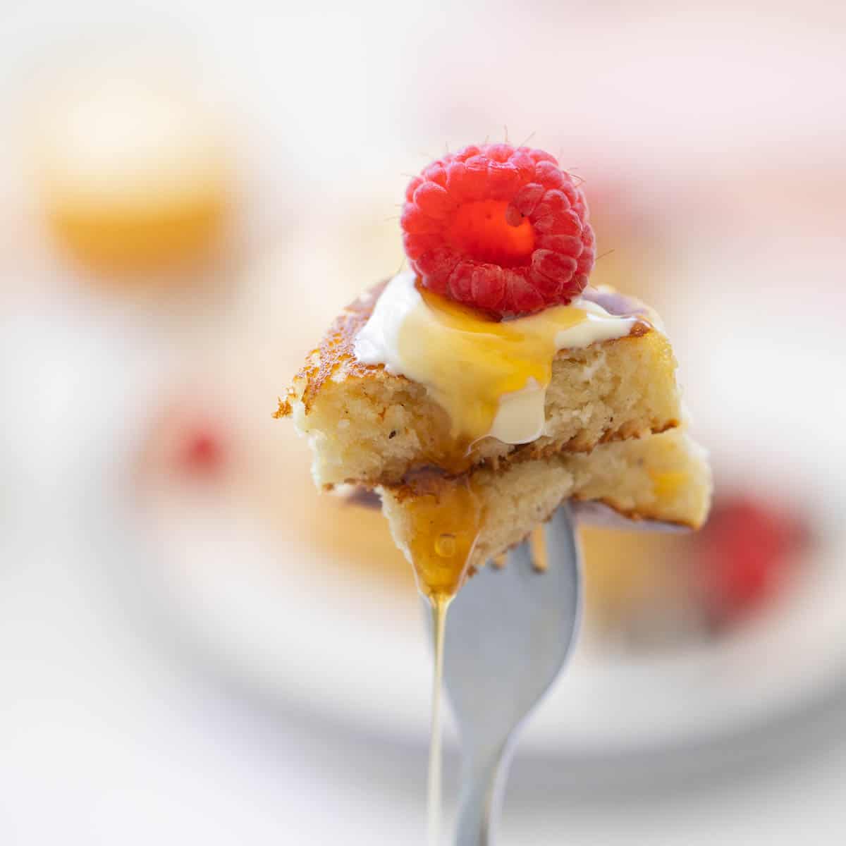Two pieces of fluffy pancakes on a fork with a raspberry, yoghurt and syrup.