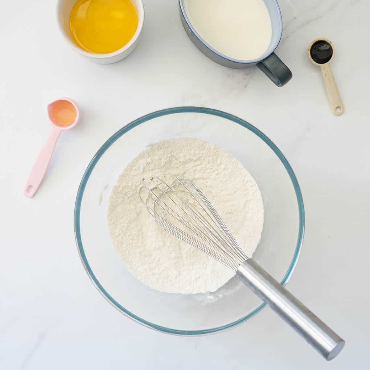 A large glass mixing bowl of dry ingredients with a whisk.