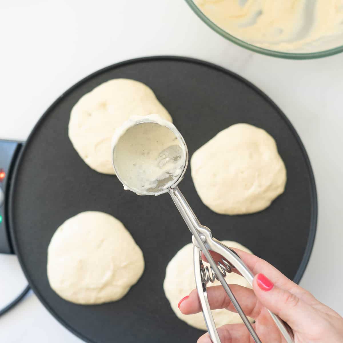 An ice cream scopp being held above, four scoops of pancake batter on a heated pancake pan.