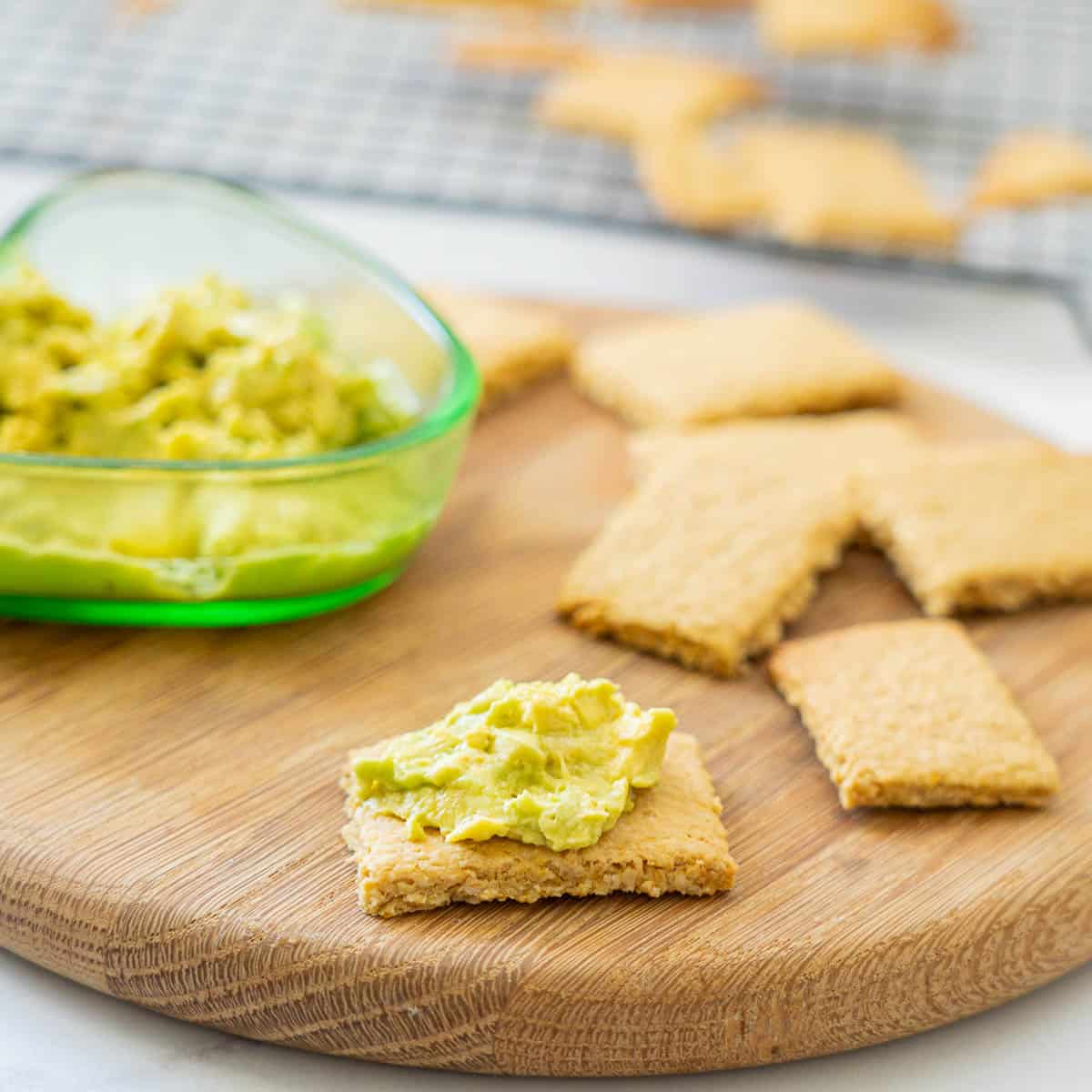 A cracker topped with avocado on a wooden board surrounded by other crackers.