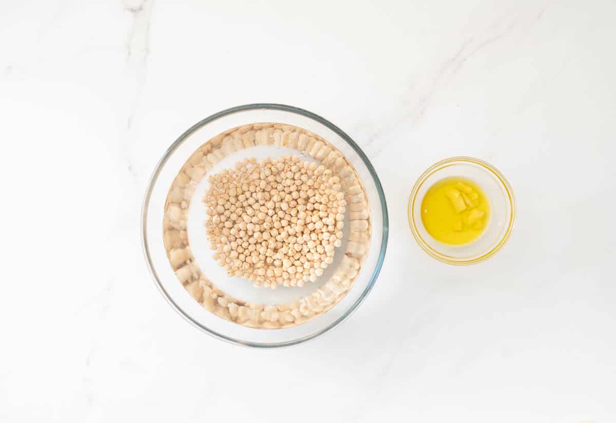 A large glass bowl of chickpeas soaking in water, next to a small bowl of garlic soaking in olive oil.