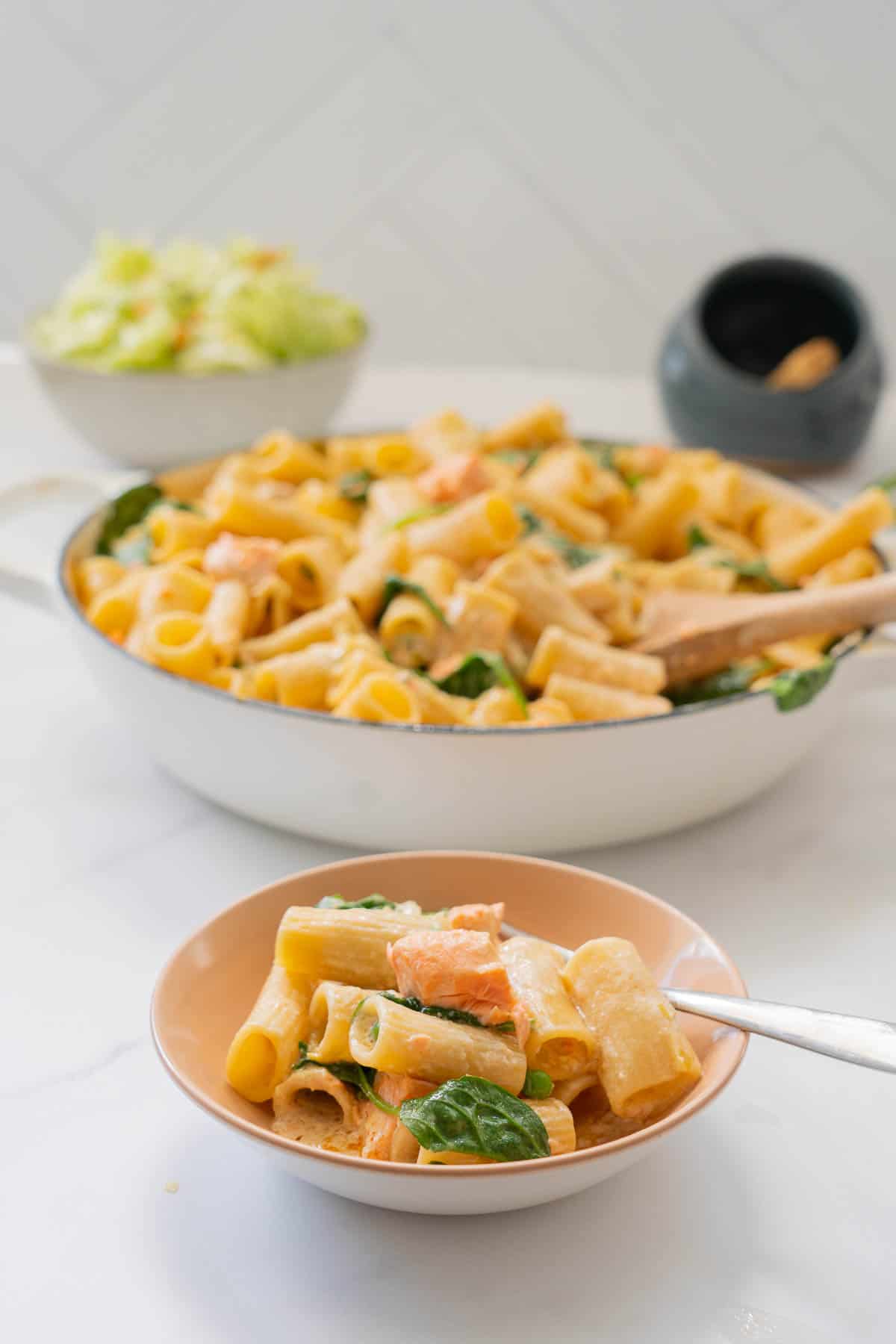 A light pink ceramic bowl cooked pasta in a cremy sauce with salmon, spinach leaves and peas also visible.