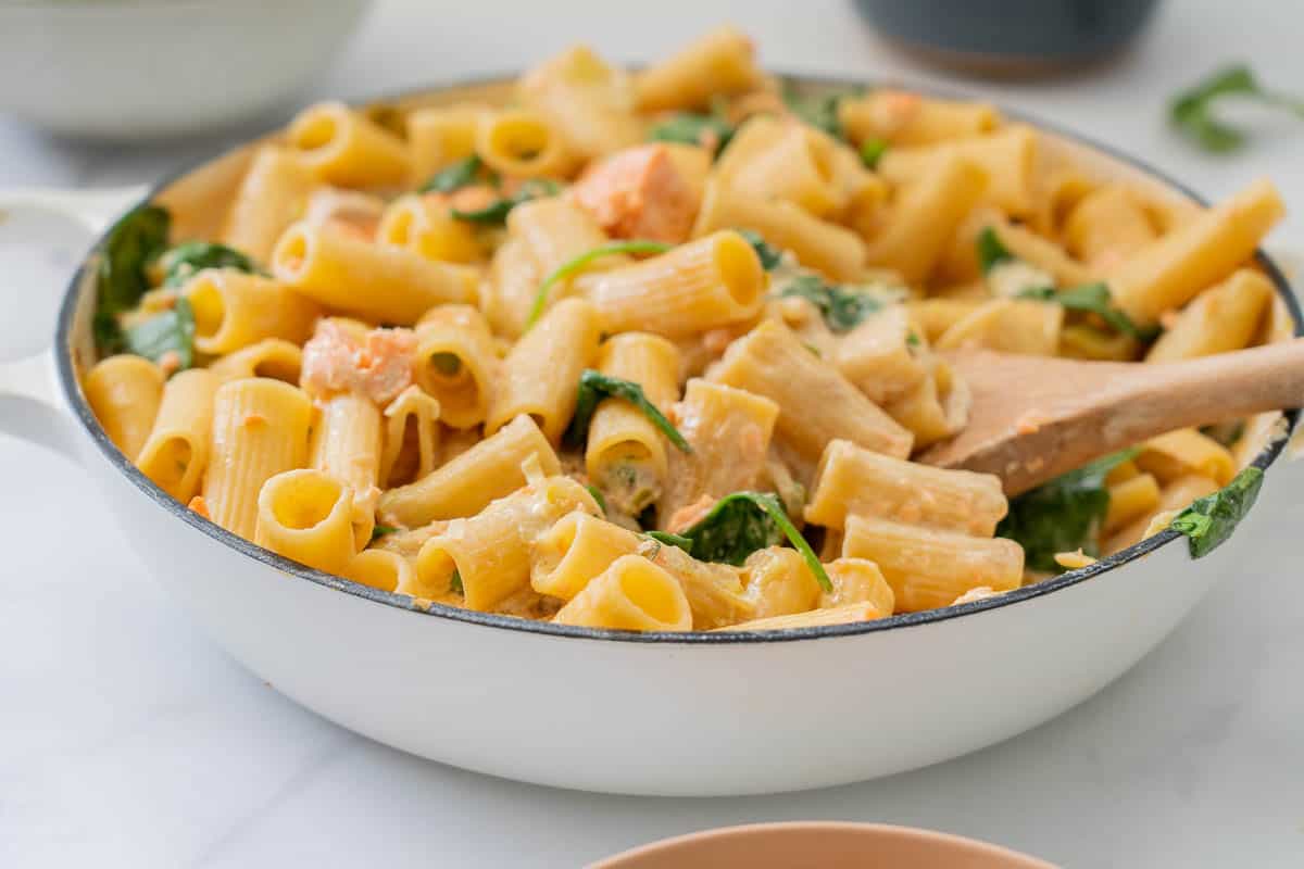 A close up of cooked pasta in a cremay sauce with salmon, spinach leaves and peas also visible.