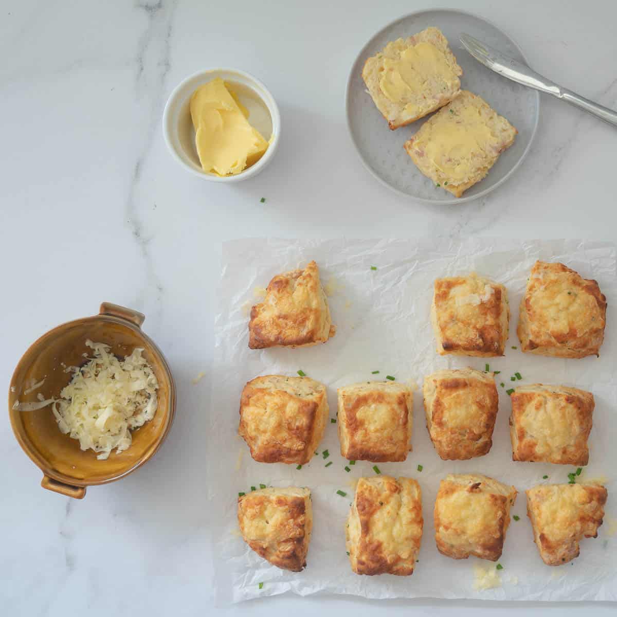 A split scone on a small ceramic plate next to eleven scones on crinkled baking paper.