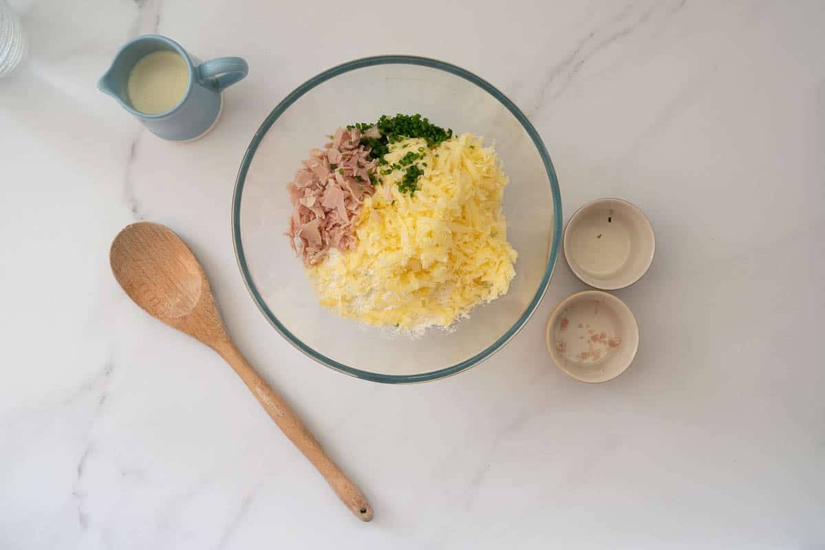 A large glass mixing bowl with, flour, grated cheese, ham and chives ready to be mixed.