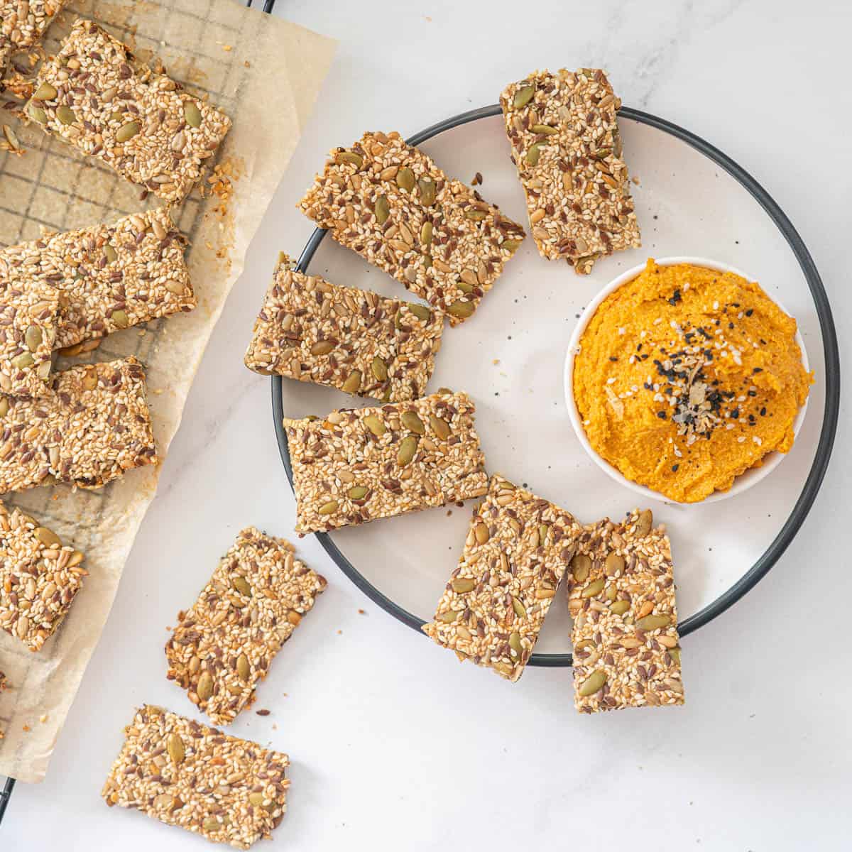 Seed crackers on a black rimmed white plate with a bowl of carrot dip.
