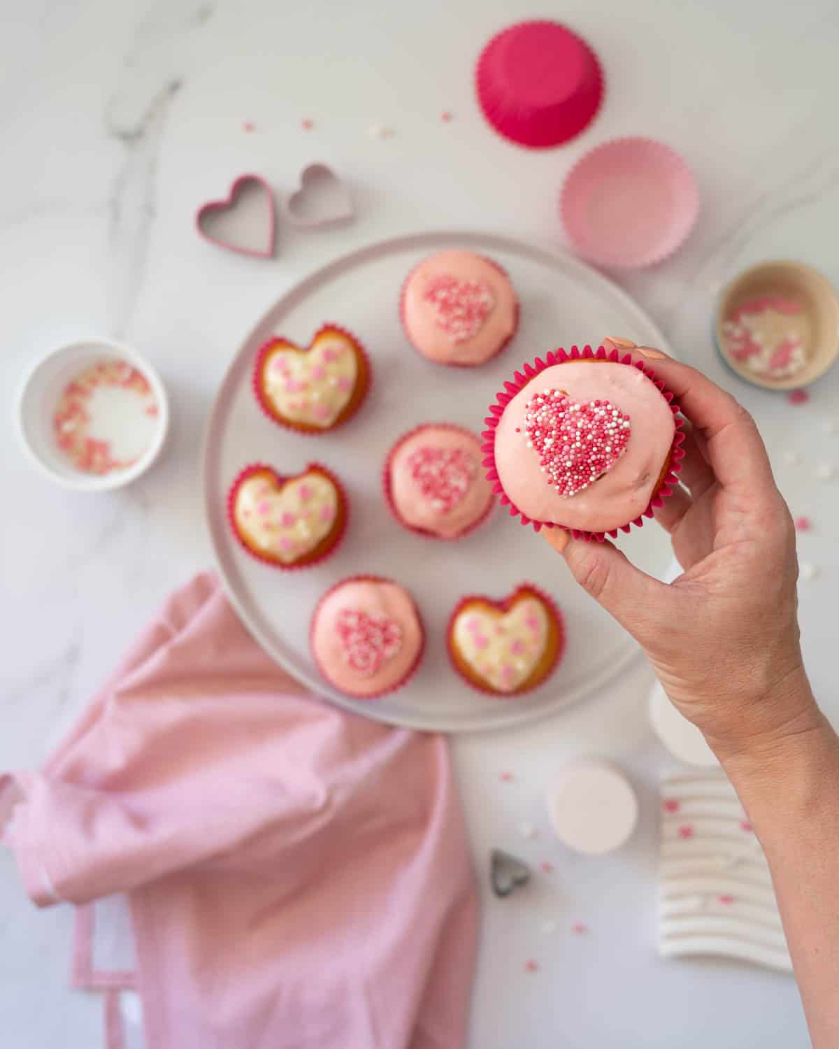 A cupcake glazed with pale pink glaze, and decorated with pink and white sprinkles in a heart shape, being held above a platter of Valentines day cupcakes.