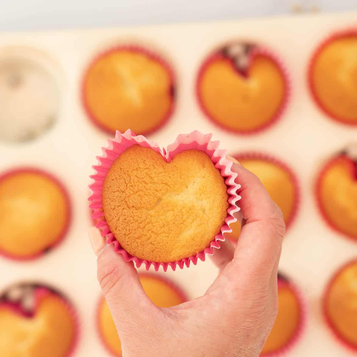 A golden brown baked heart shaped cupcake being held above a tray of cupcakes.