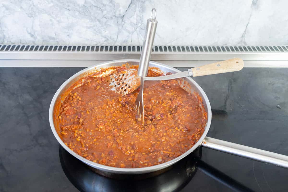 A potato masher in a Vegetarian bean nacho mix simmering in a stainless steel fry pan.