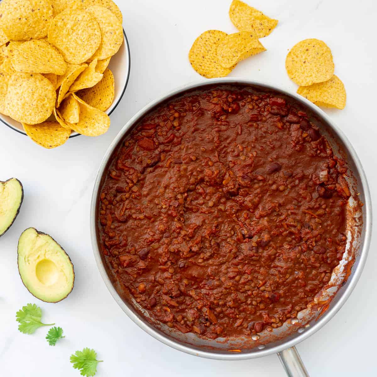 A large stainless stell fry pan of bean based nacho mix on a table top with corn chips, avocado and coriander leaves.