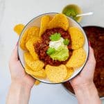 A bowl of bean nachos with corn chips, sourcream and mashed avocado being held in two hands.