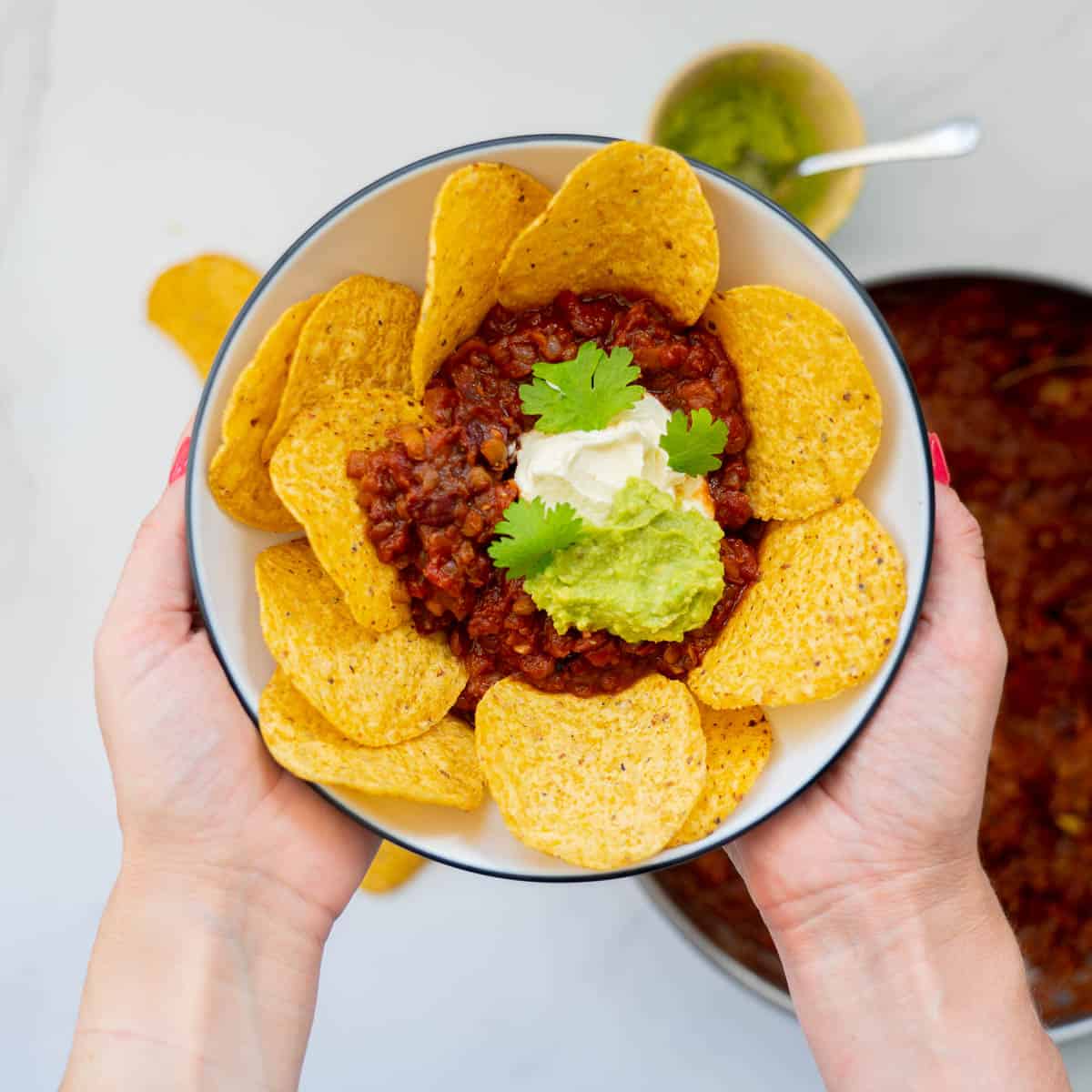 A bowl of bean nachos with corn chips, sourcream and mashed avocado being held in two hands.