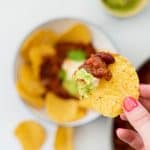 A corn chip loaded with bean based nacho mix, avocado and sour cream being held above a dinner table.