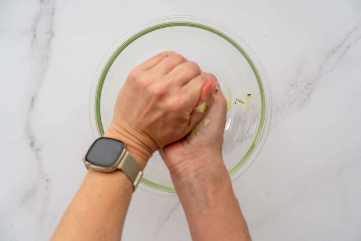 Grated zucchini being squeezed above a glass bowl that is collecting the green watery liquid.