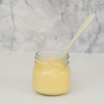 A small glass jar of apple puree with a green bamboo spoon in front of a marble splash back.