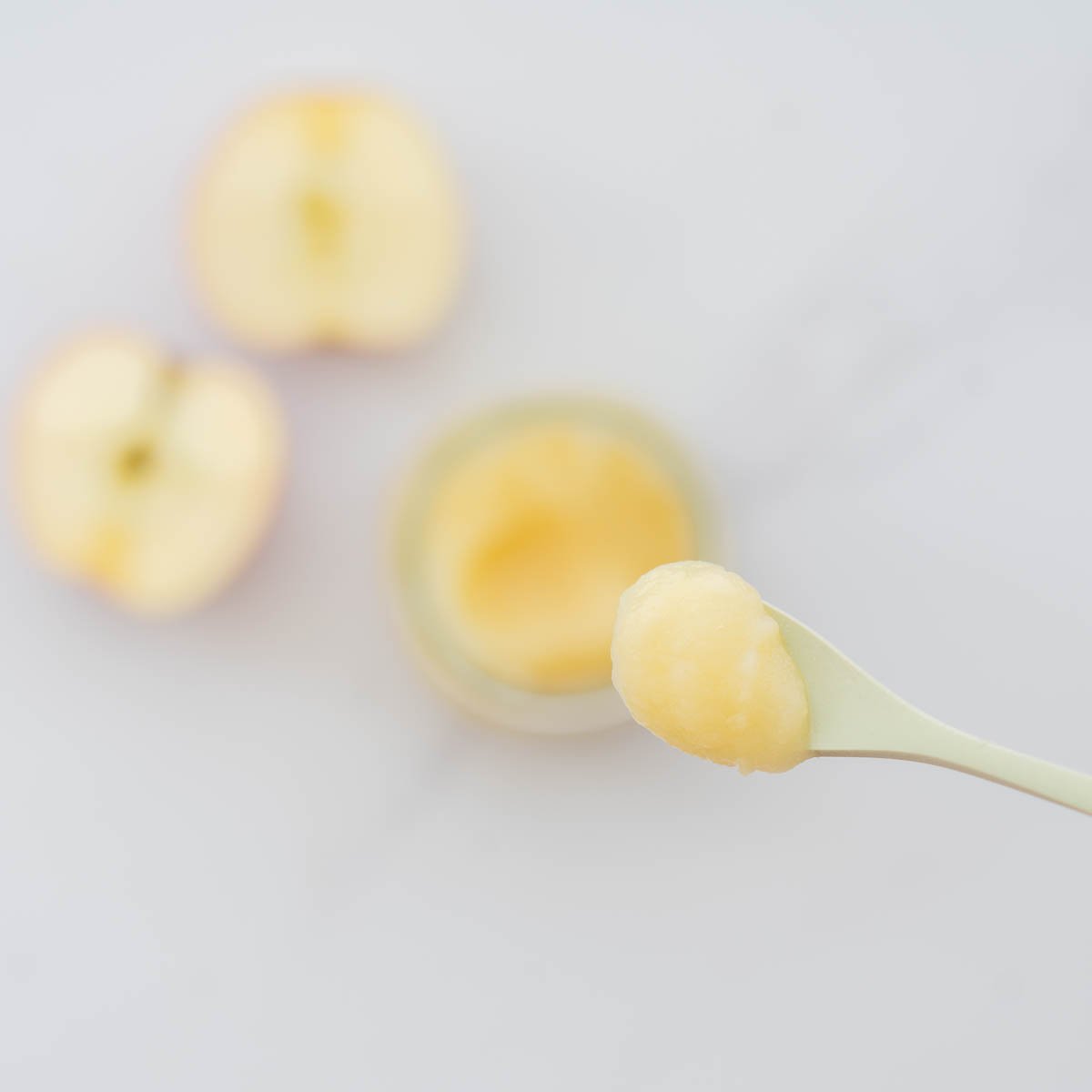 A spoonful of apple puree being held above a jar of apple puree.