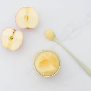 Apple puree in a small glass jar, with a spoonful of puree and halved apples on the bench next to the jar.