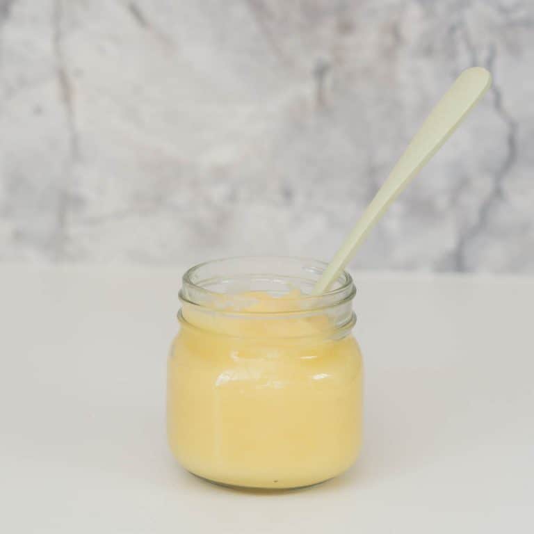 A small glass jar of apple puree with a green bamboo spoon in front of a marble splash back.