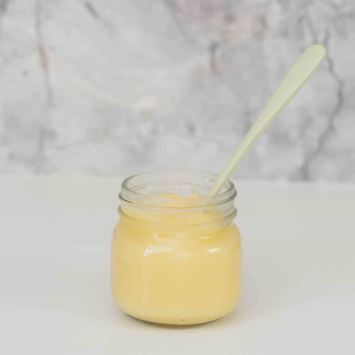 A small glass jar of apple puree with a green bamboo spoon in front of a marble splash back.