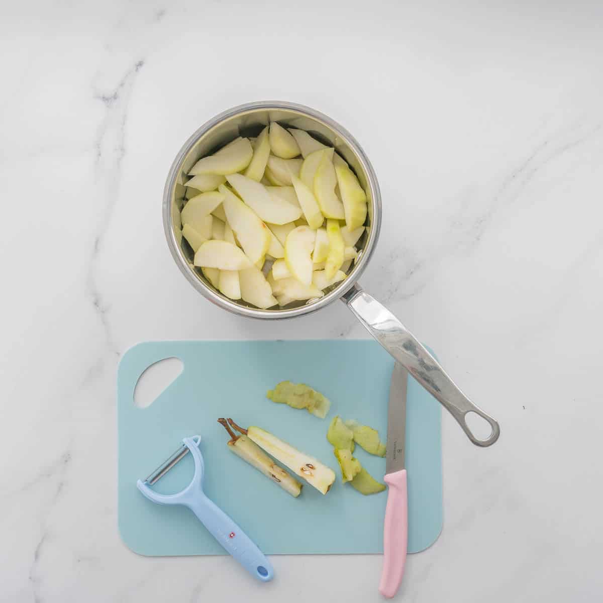 A stainless steel saucepan of peeled and sliced peers next to a chopping board with pear offcuts.