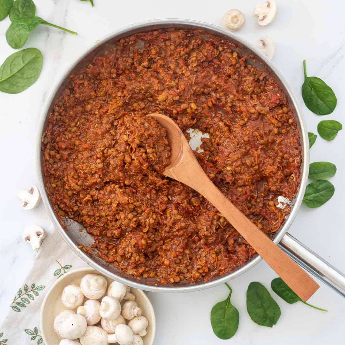 A large fry pan of vegan bolognese on a table top scattered with spinach leaves and button mushrooms.