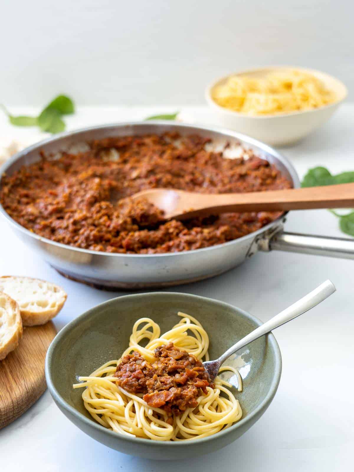 A green ceramic bowl of spaghetti bolognese on a table in front of a large fry pan of bolognese sauce.