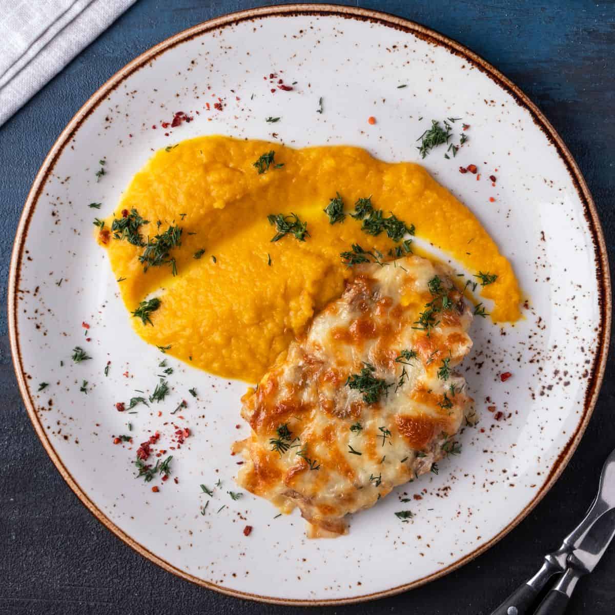 A grilled piece of chicken on a white plate with pureed carrot, scattered with herbs.