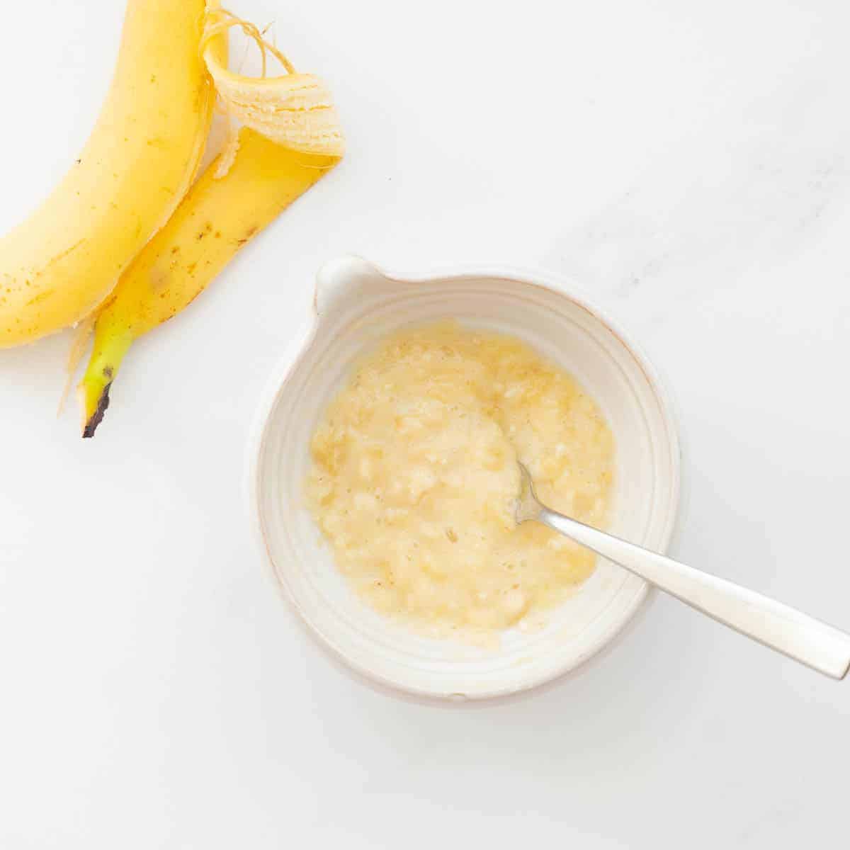 Banana skin next to a white bowl that has mashed up banana with a fork