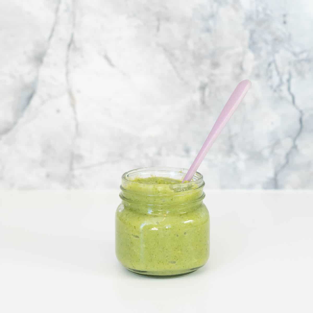 A glass jar of green puree sitting in front of a grey marble splashback.