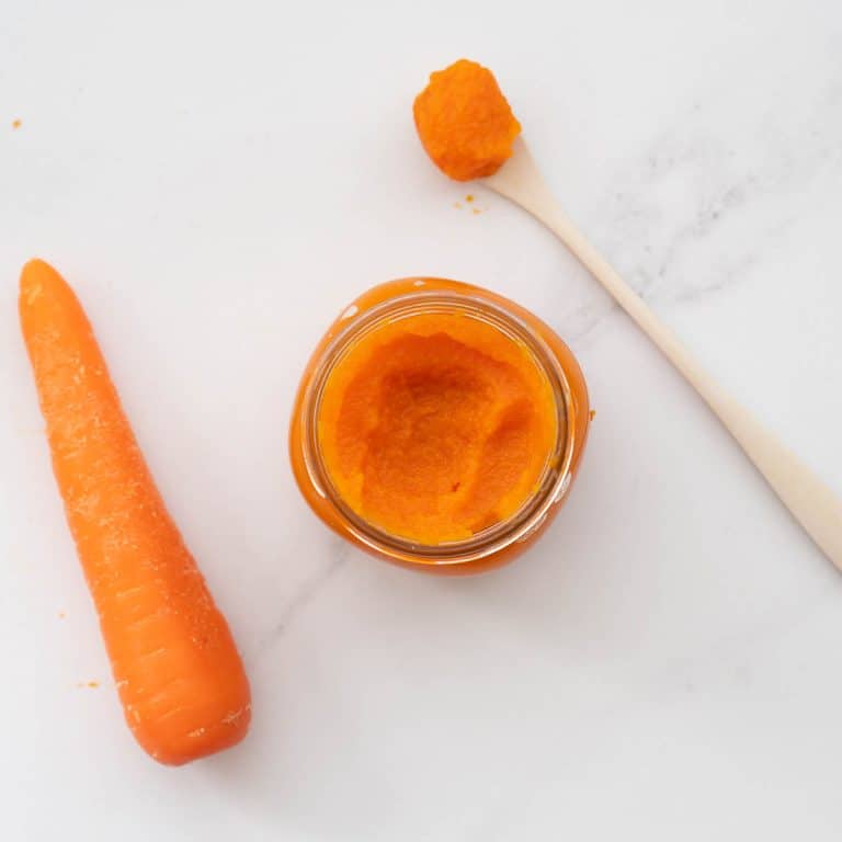 A jar of orange carrot puree on a bench next to a carrot and a bamboo spoon loaded with carrot puree.
