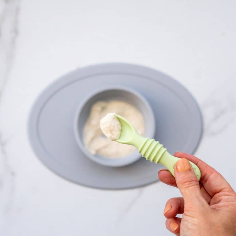 A spoonful of chicken puree being held above a grey baby plate of chicken baby food.