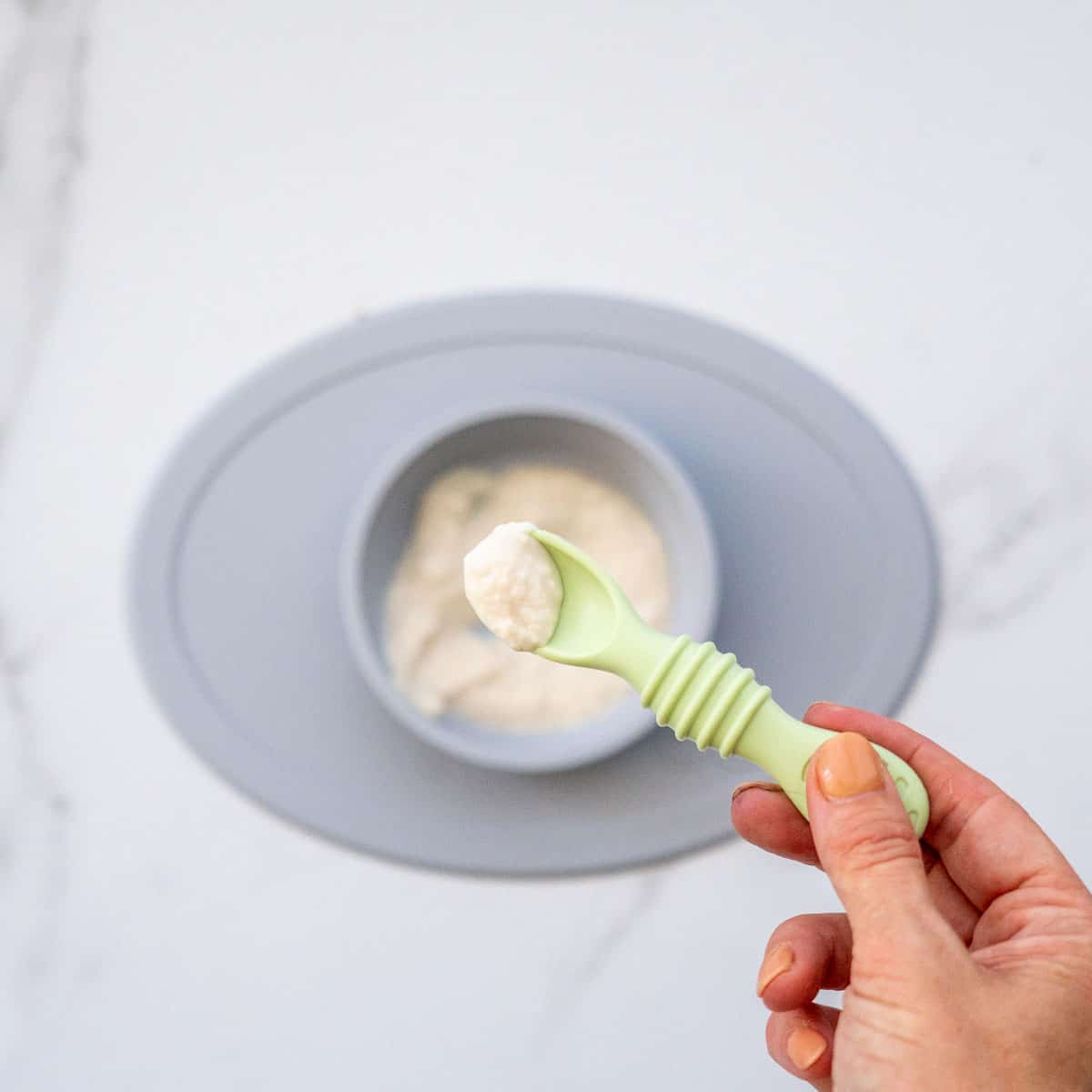 A spoonful of chicken puree being held above a grey baby plate of chicken baby food.