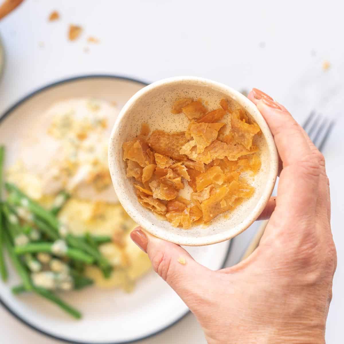 A bowl of small pieces of crispy chicken skin being held above a meal of poached chicken.