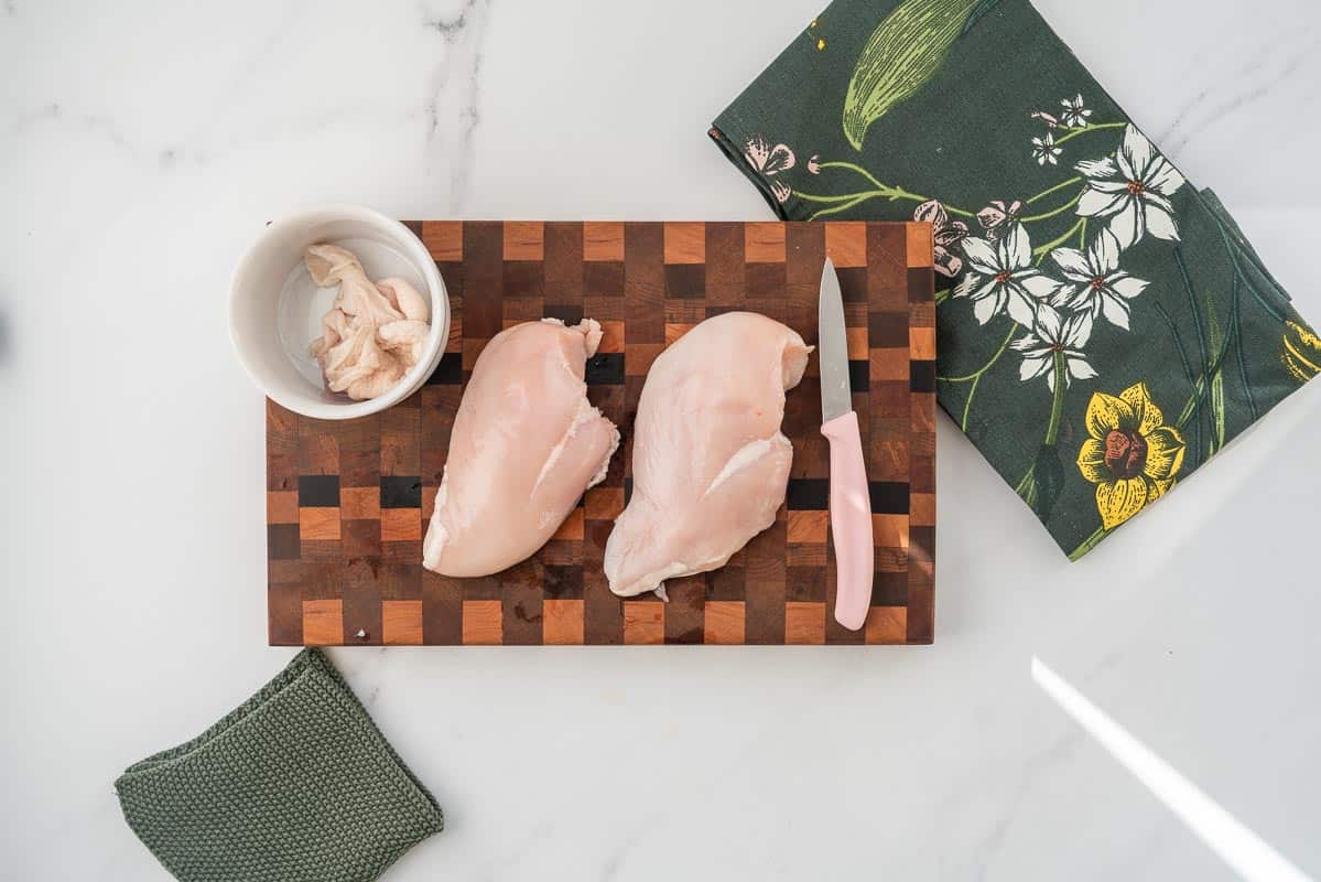 2 Chicken breasts on a wooden butcher block, skin removed and separated into a small ceramic ramekin.