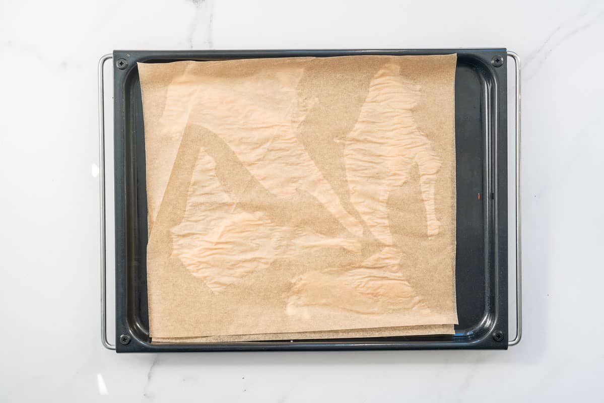 Four pieces of thinly stretched chicken skin on a baking paper lined tray, topped with a second piece of baking paper.