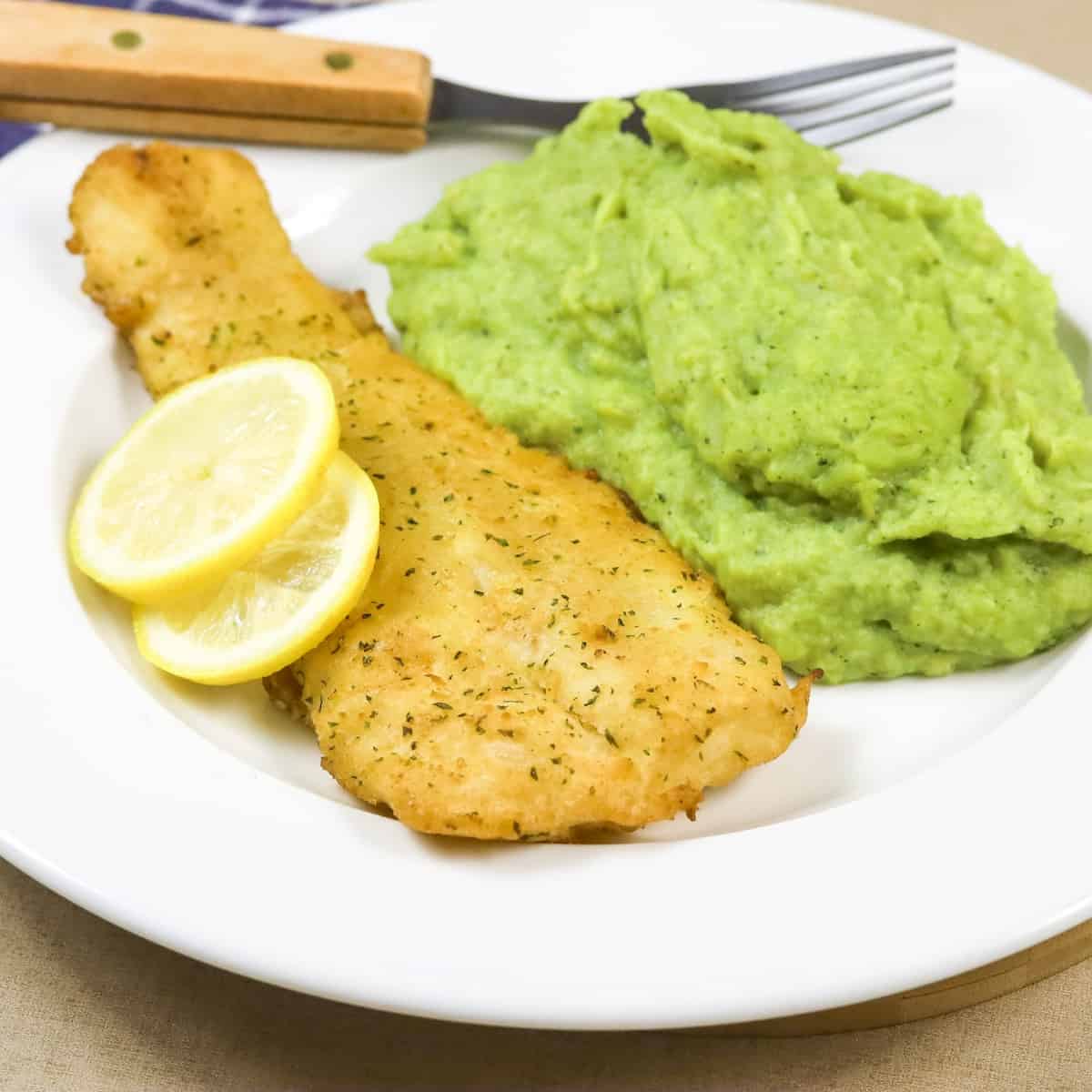 A white dinner plate with a piece of crumbed fish, broccoli puree and lemon slices.