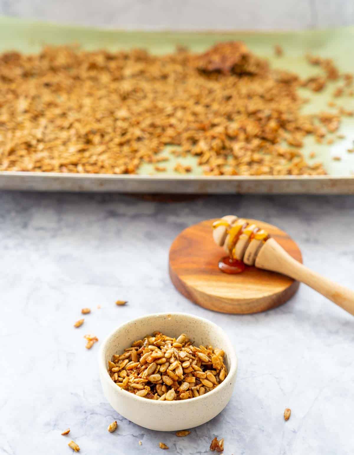 Faded in the background the over tray with the roasted sunflower seeds and the honey dipper stick on the wooden coaster.In front is the oatmeal bowl with the roasted seeds
