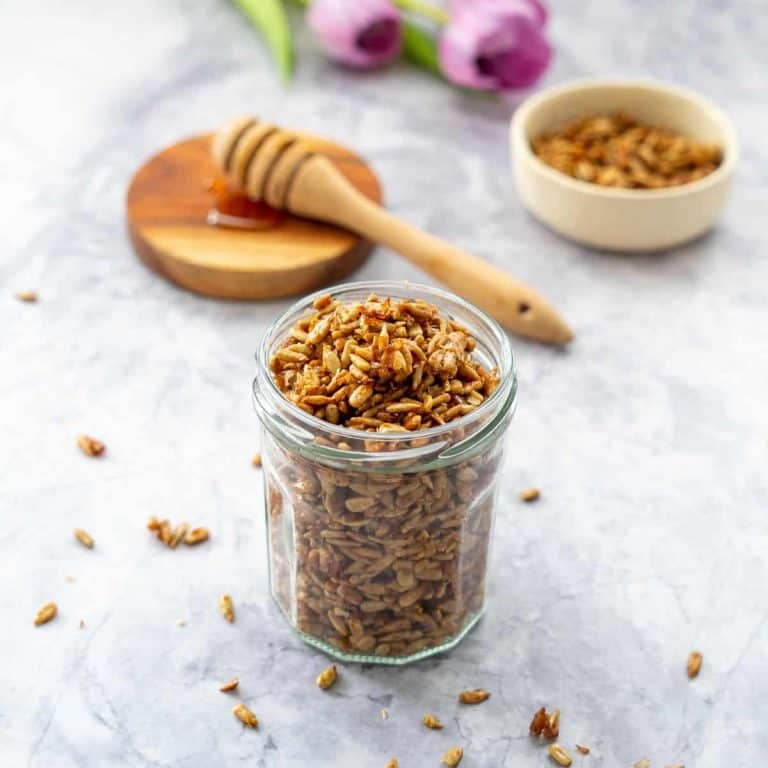 A glass jar of roasted sunflower seeds, with a spoonful of honey and purple tulips in the background.