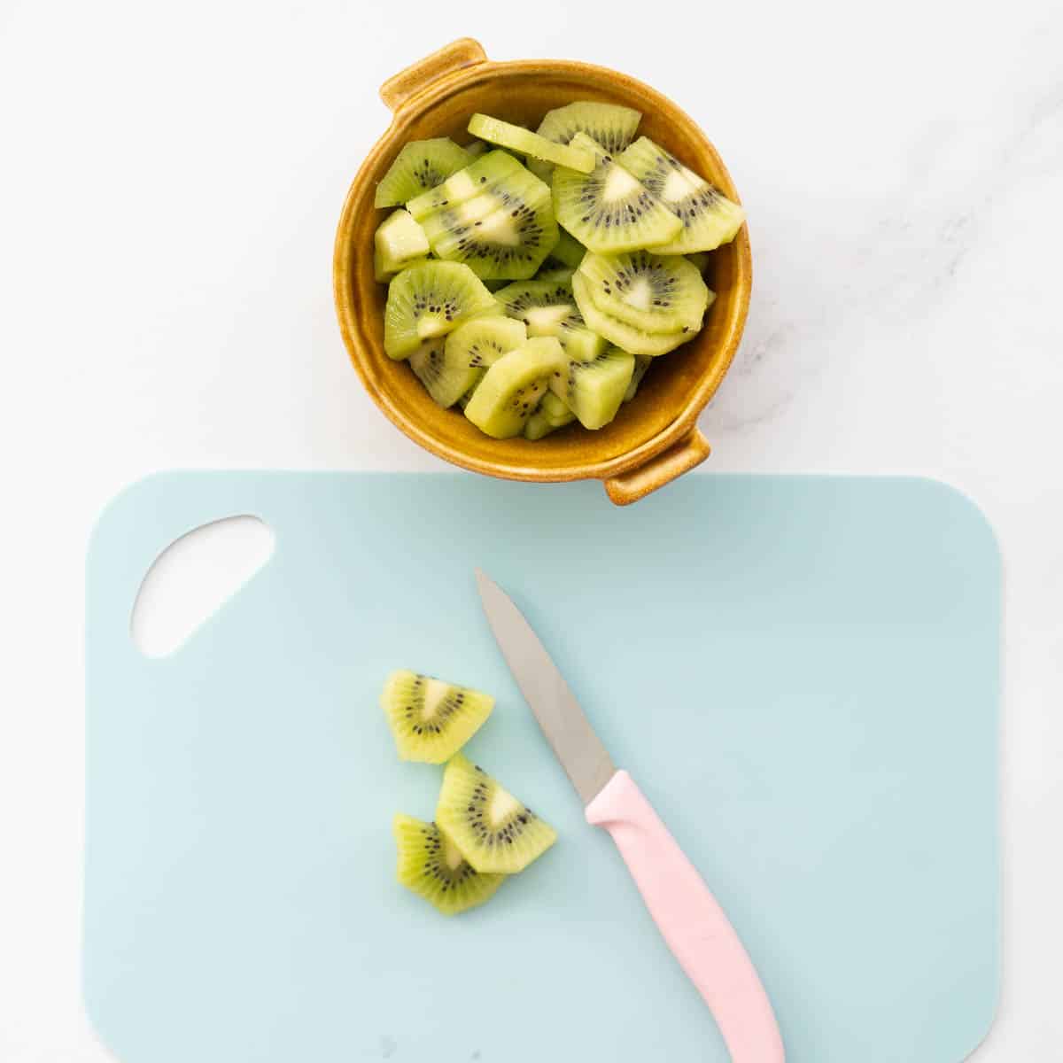 Peeled and sliced kiwifruit in a ceramic bowl, next to a light blue chopping board and pink handled fruit knife.