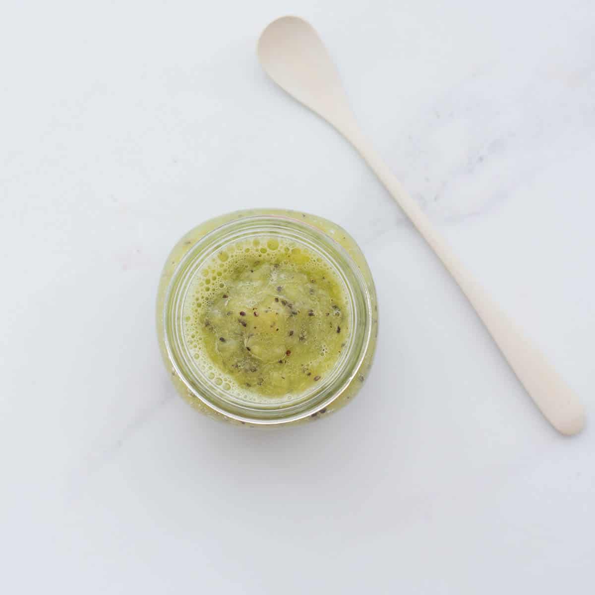A small glass jar of kiwi puree on a bench top with a cream bamboo spoon next to it.