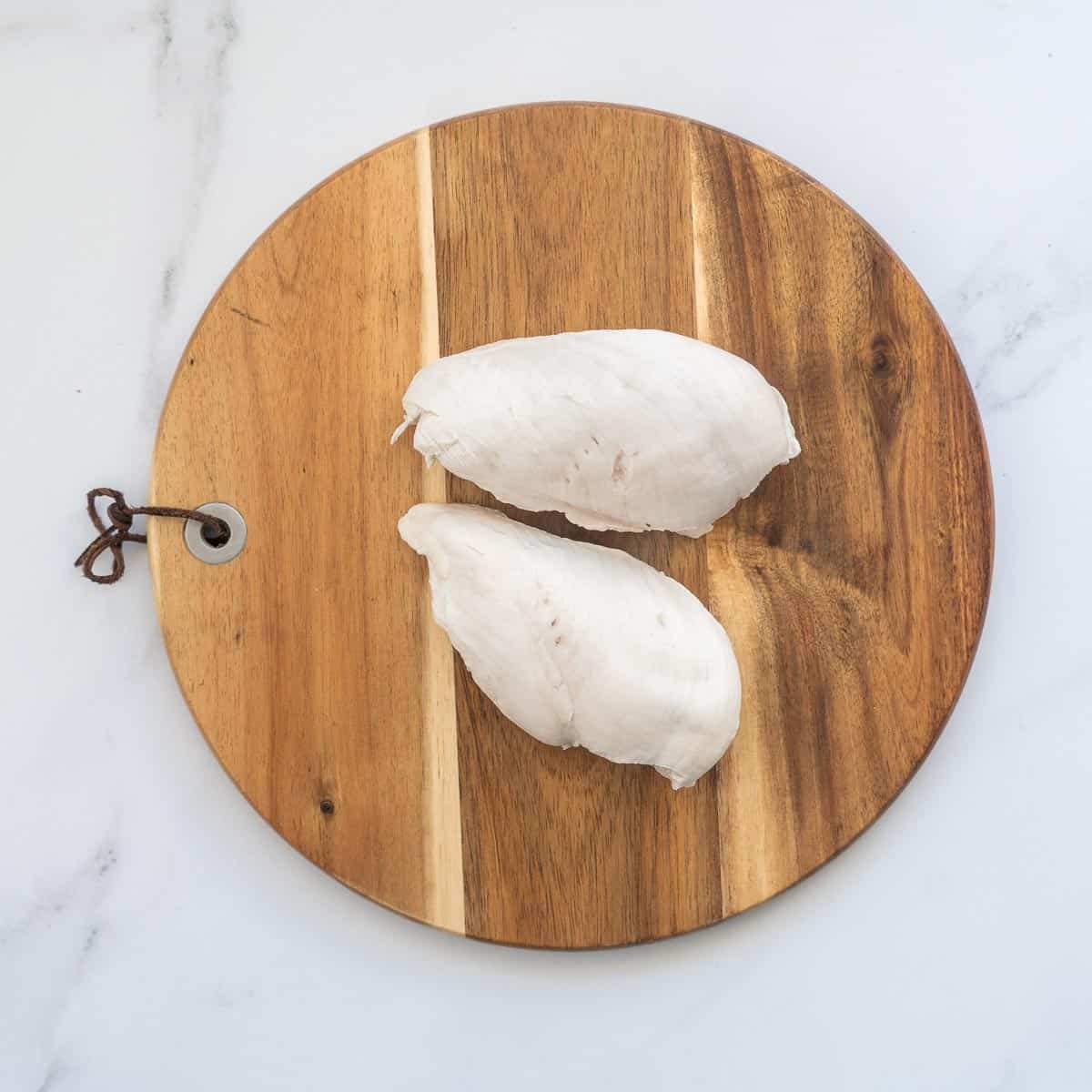 Two cooked chicken breasts resting on a round wooden chopping board.