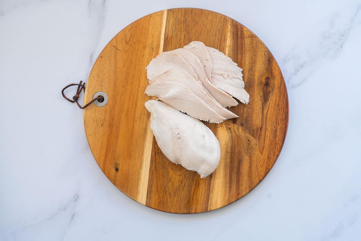 One whole poached chiken breast and one sliced chicken breast on a round wooden chopping board.