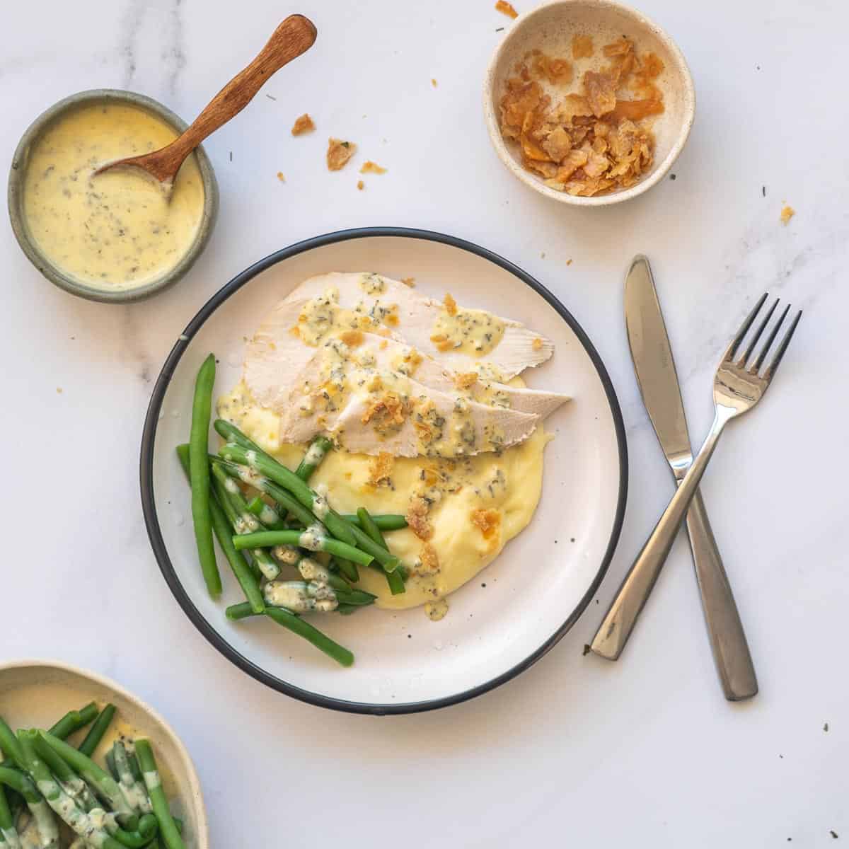 Slices of chicken breast on mashed potatoes with green beans and a yellow sauce with sides of green beans, sauce and crispy chicken skin in bowls.