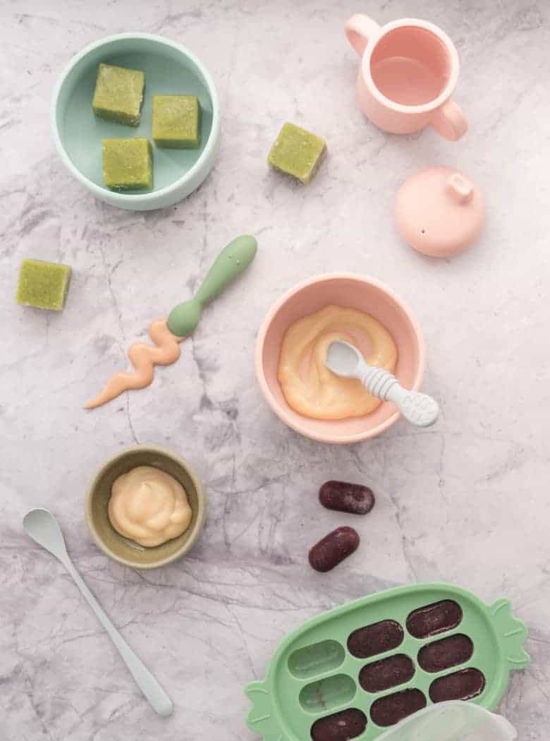 Baby purees in different forms, in serving bowls, on baby spoons, in freezer trays, smeared on marble bench top.