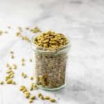 A jar of roasted pepitas on a marble bench, some pepitas are scattered on the bench top.