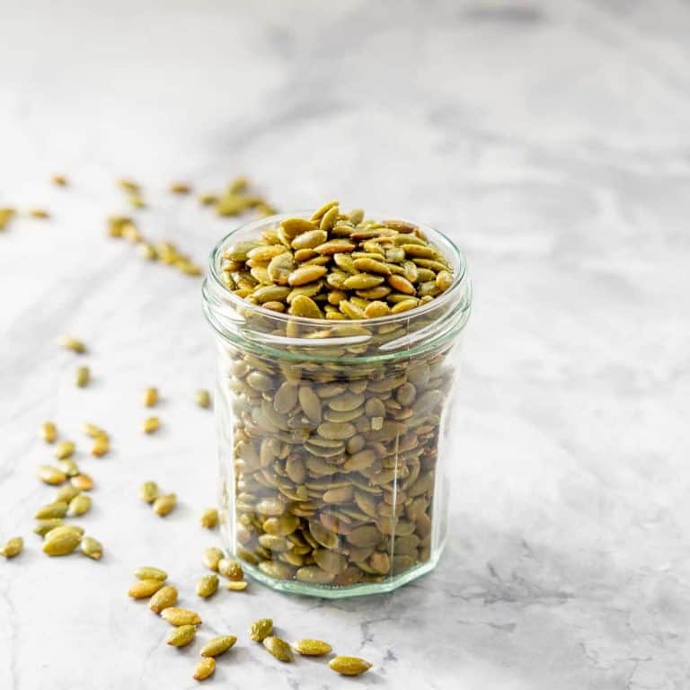 A jar of roasted pepitas on a marble bench, some pepitas are scattered on the bench top.